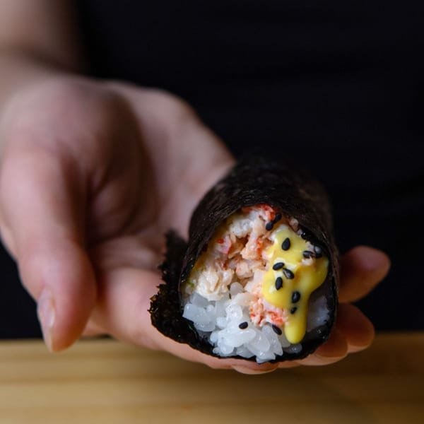 A hand holds a Temakase hand roll filled with rice, crab, and yellow sauce, topped with black sesame seeds.