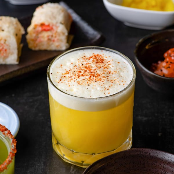 A yellow cocktail with foamy top and red spice sits among sushi rolls and small dishes.