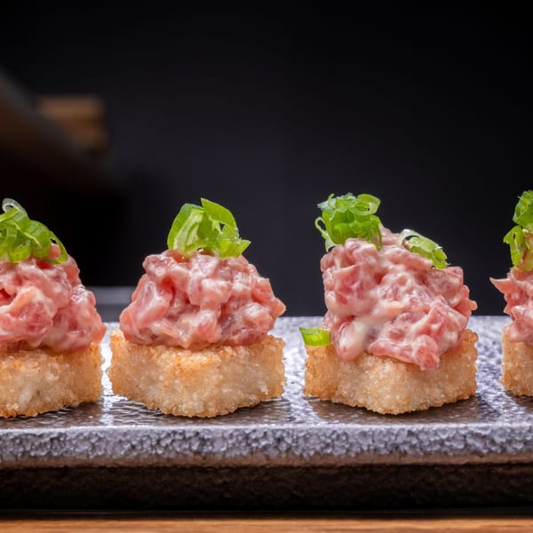 Four pieces of Temakase spicy tuna crispy rice, topped with green onions, on a dark stone plate.