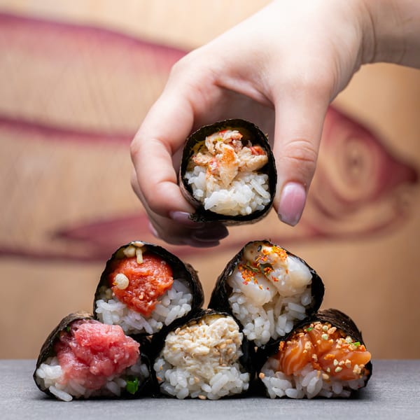 A hand holding a crab hand roll above a pyramid of five other Temakase sushi hand rolls.