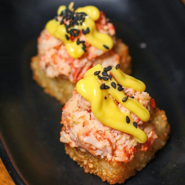 Two crispy rice sushi bites topped with crab, yellow sauce, and black sesame seeds.