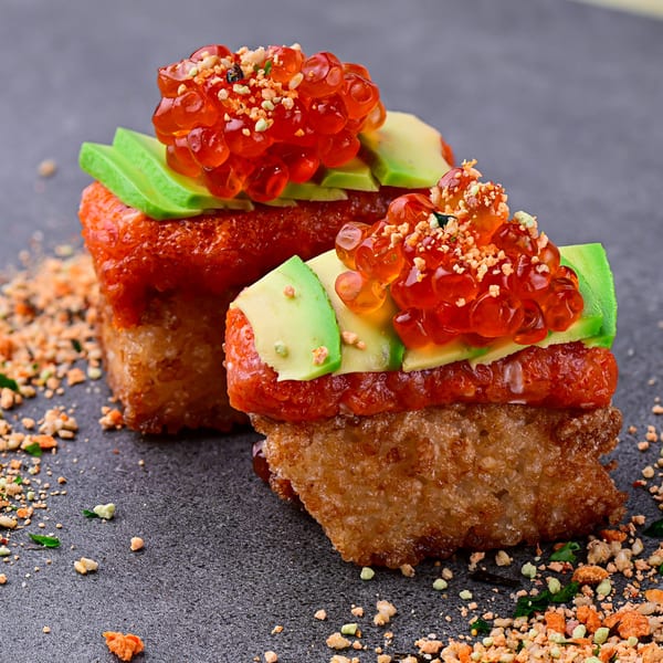 Two crispy rice sushi pieces topped with avocado, salmon roe, and crushed nuts on a dark surface.