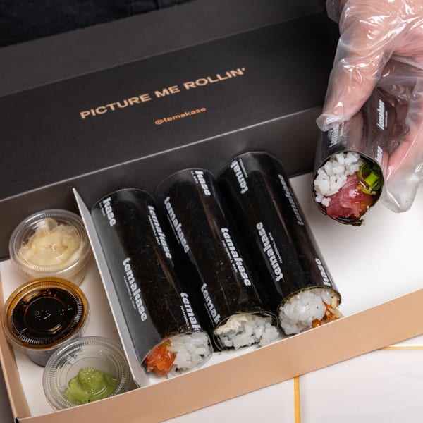 A hand in a plastic glove holds a Temakase hand roll over a box of three more rolls and sauces.