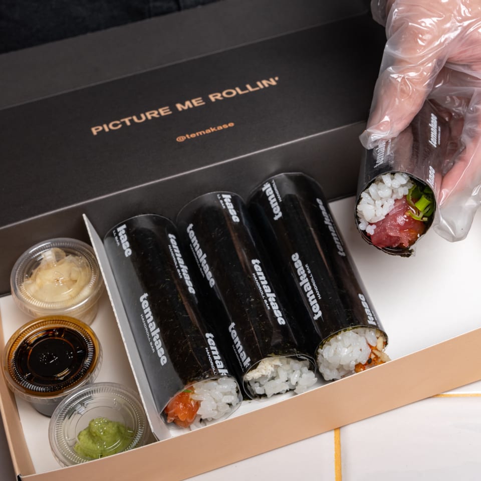 A hand in a plastic glove holds a Temakase hand roll over a box of three more rolls and sauces.