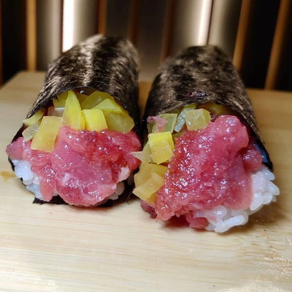 Two Temakase hand rolls with minced tuna, rice, and yellow pickled daikon on a wooden board.