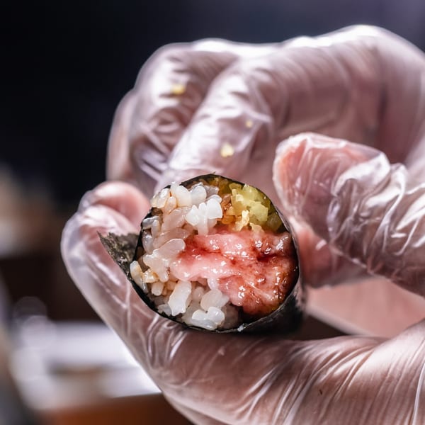 Gloved hands holding a Temakase hand roll, filled with rice, tuna, and yellow pickled daikon.