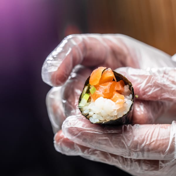Gloved hands holding a freshly prepared Temakase hand roll filled with salmon, rice, and green onions.