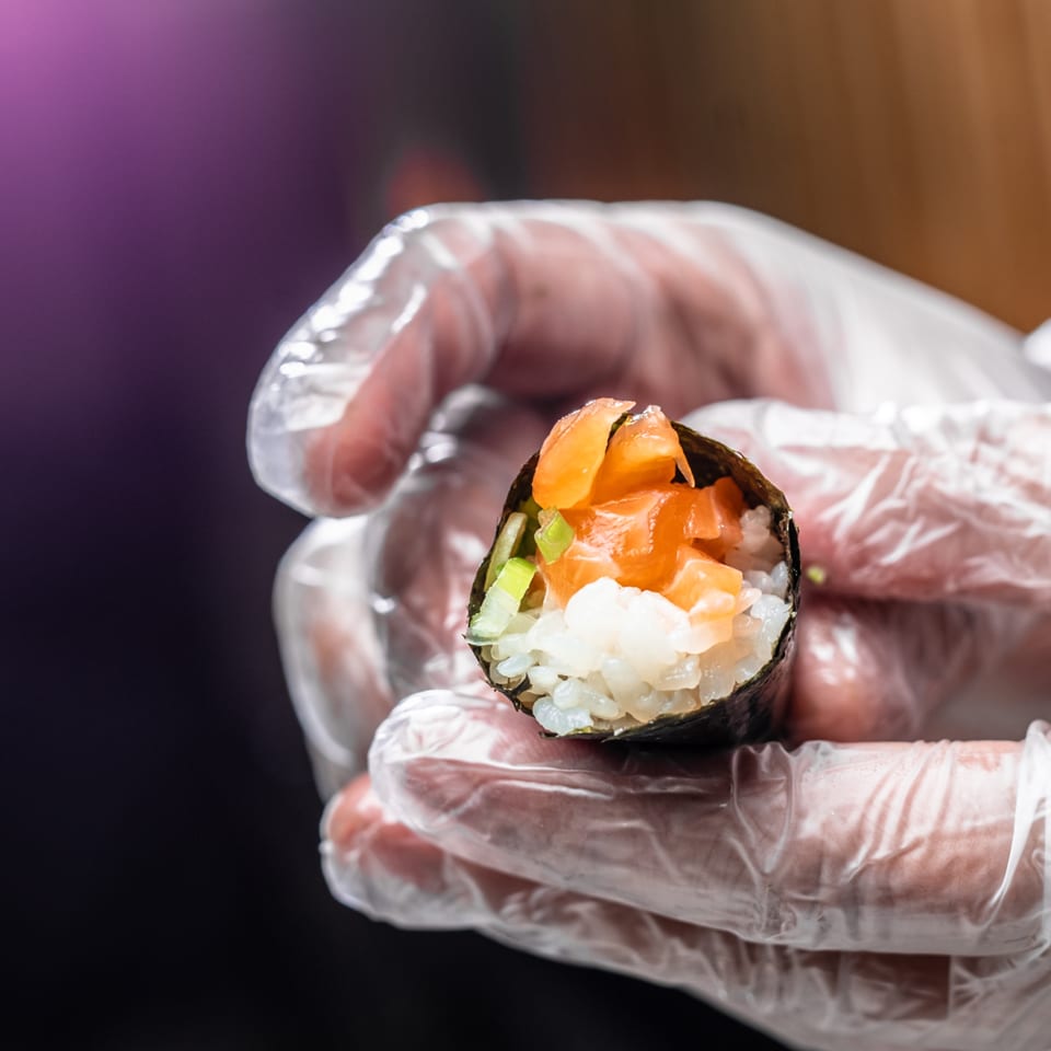 Gloved hands holding a freshly prepared Temakase hand roll filled with salmon, rice, and green onions.