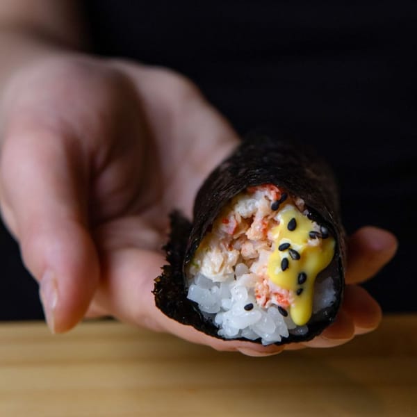 A hand holds a Temakase hand roll filled with rice, crab, yellow sauce, and black sesame seeds.