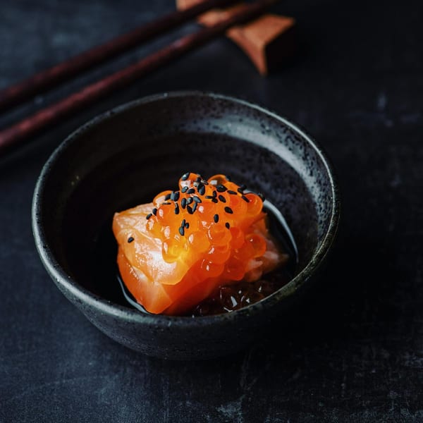 Close-up of salmon sashimi topped with bright orange ikura and black sesame seeds in a dark bowl.