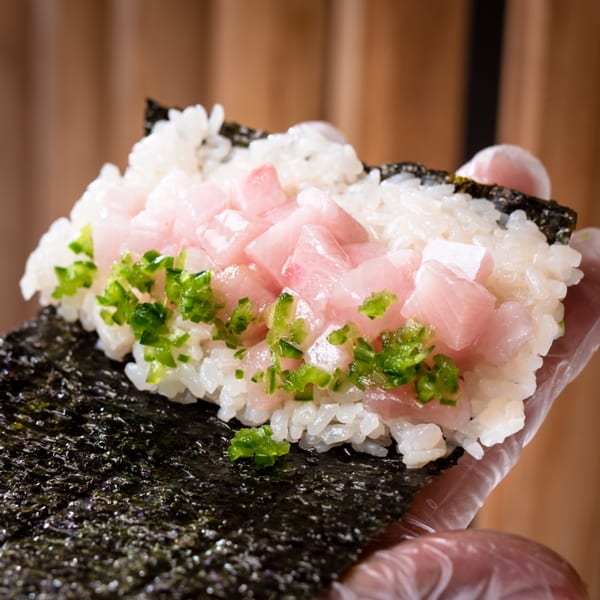 A hand in a clear glove holds a Temakase hand roll with rice, diced tuna, and green onions.