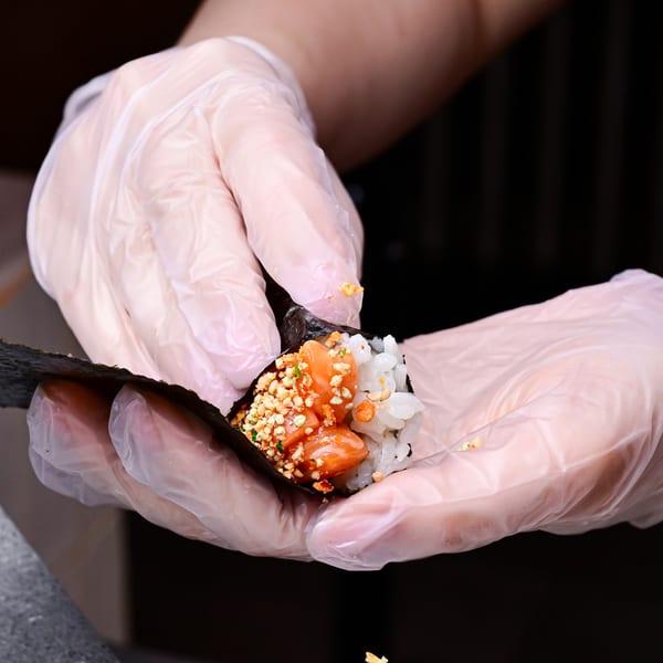 Gloved hands preparing a Temakase hand roll with salmon, rice, and crunchy toppings in nori.