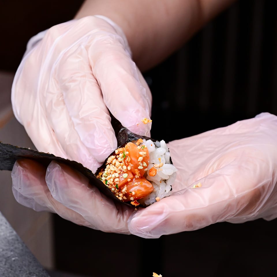 Gloved hands preparing a Temakase hand roll with salmon, rice, and crunchy toppings in nori.
